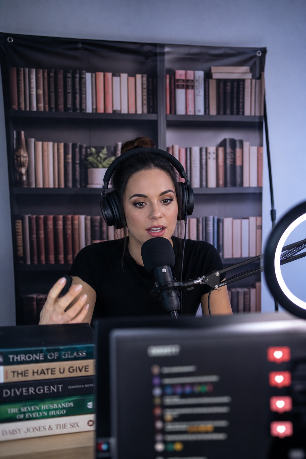 Streaming setup with a young content creator wearing studio headphones, a condenser microphone on a boom arm and a ring light. Behind her, a printed fabric backdrop reproduces the spines of a curated bookshelf in muted pinks and beiges. In the foreground, a stacked pile of contemporary BookTok bestsellers — Throne of Glass, The Hate U Give, Divergent, The Seven Husbands of Evelyn Hugo, Daisy Jones & The Six — frames the shot. A monitor on the right displays a live-stream chat with floating heart reactions.